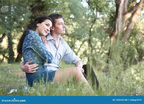 Young Couple Sitting On The Grass In The Forest And Looking On Sunset