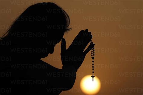 Silhouette Of Faithful Woman Praying With Rosary Beads At Sunset As