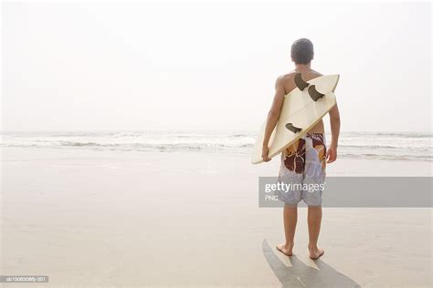 Teenage Boy Standing With Surfboard On Beach Facing Ocean Rear View