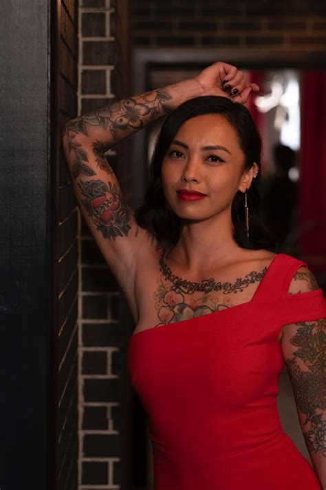 A Woman In A Red Dress With Tattoos On Her Arms And Shoulder Leaning Against A Brick Wall