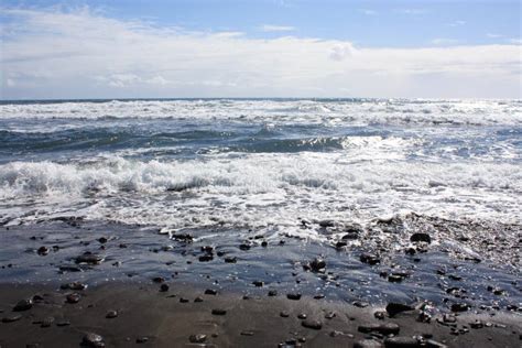 Waves Rolling Onto A Black Sand Pebble Strewn Beach On A Cloudy Day In