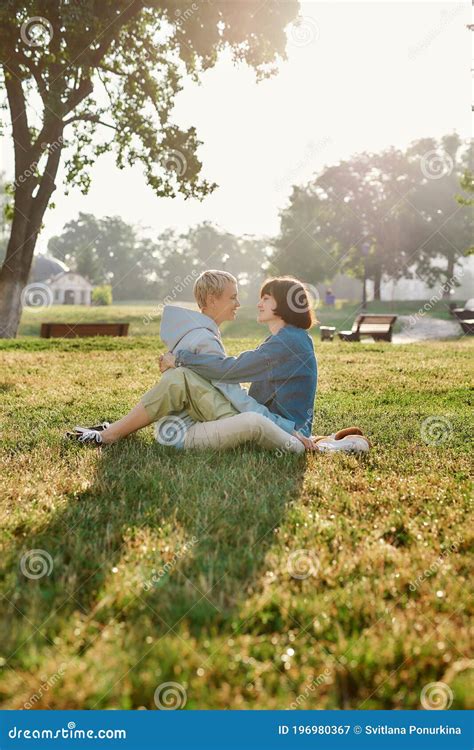 happy lesbian couple of women looking at each other while sitting on the grass in summer park
