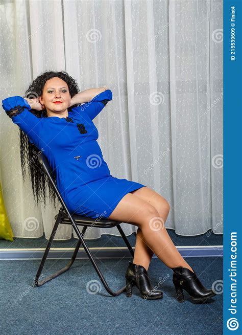 A Curly Haired Brunette In Blue Sits On A Chair And Puts Her Hands Behind Her Head Stock Image