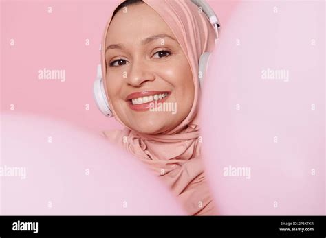 middle aged beautiful muslim woman in headphones and pink hijab smiling peeking out from