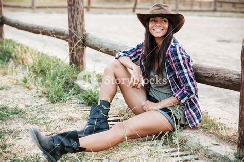 Smiling Happy Cowgirl In Hat Sitting And Resting At The Ranch Fence