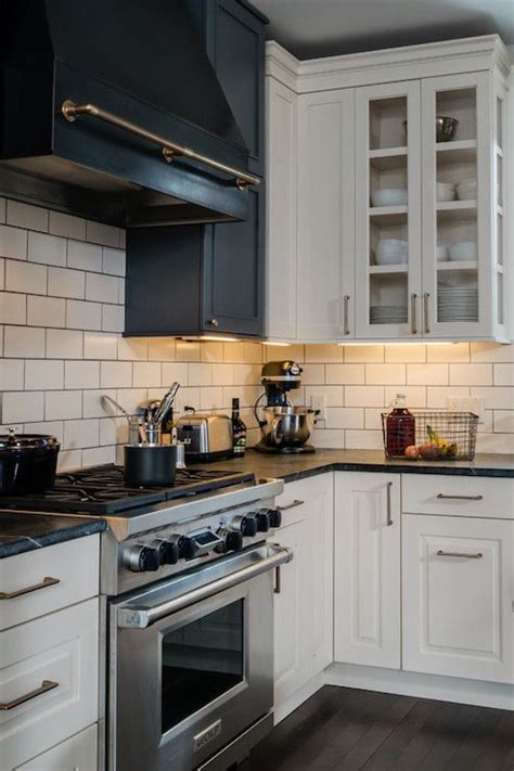 Mounted to marble mini brick backsplash tiles, a black range hood accented with brass. Beautiful kitchen with white raised panel cabinets paired with brass hardware and soapstone ...
