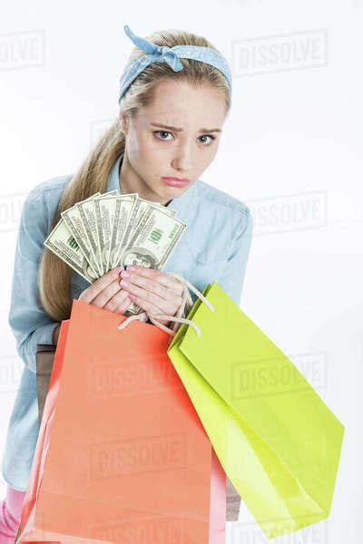 Sad Young Woman Holding Shopping Bags And Dollar Banknotes On White