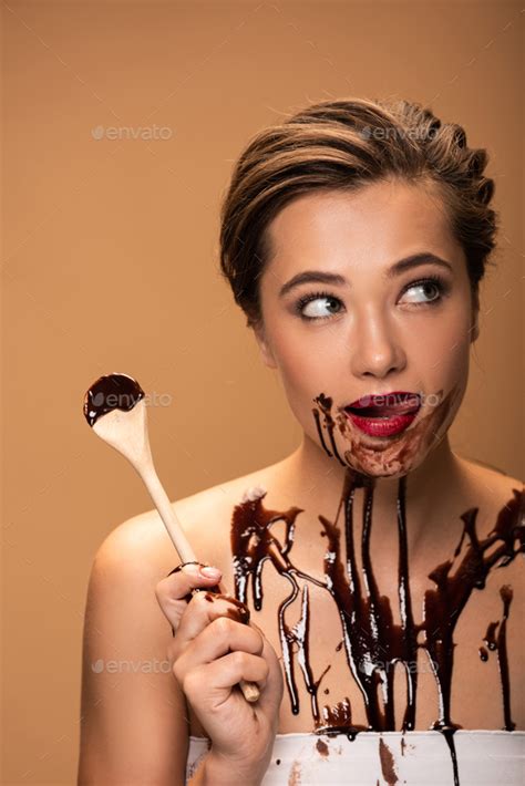 beautiful woman licking red lips holding wooden spoon and looking away with chocolate spills on