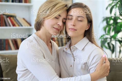 beautiful middle aged mom and her adult daughter are hugging and smiling while sitting on couch