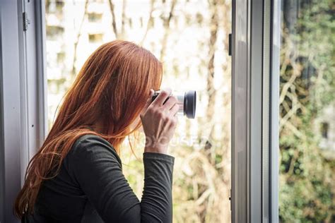Redheaded Woman Leaning Out Of Window Taking Photo With Camera — People
