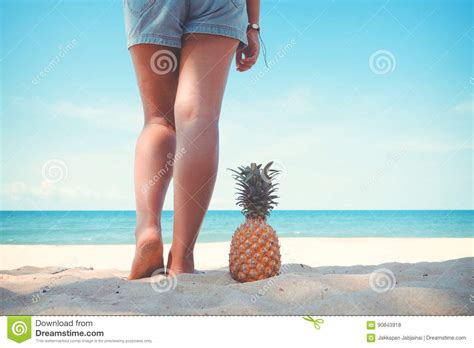 Tanned Legs Of Young Woman Standing With Pineapple At Tropical Beach In