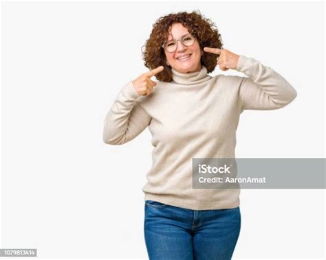 아름 다운 중간 어른 고위 여자 웃 고 자신감 보여주는 손가락 치아와 입 지적 고립 된 배경 위에 터틀 넥 스웨터와 안경 착용 건강 개념입니다 가리키기에 대한 스톡 사진 및