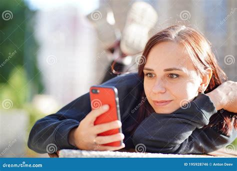 Young Woman Lying On Her Stomach Outside On A Bench With Her Legs Up And Browsing Her Cell
