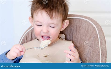 A Happy Child Eats Porridge With A Spoon While Sitting On A High Chair