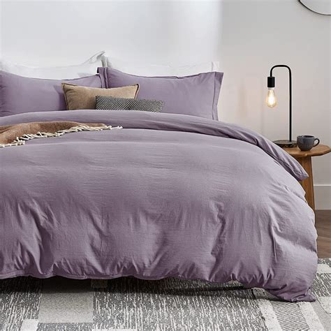 A Bed With Purple Comforter And Pillows In A White Room Next To A Lamp