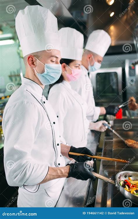 Chefs In Protective Masks And Gloves Prepare Food In The Kitchen Of A