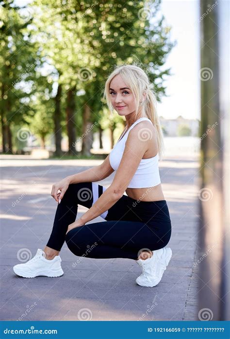Young Fit Blond Woman Wearing White Top And Black Leggings Squatting