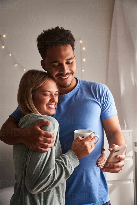 Side View Of Smiling Multiracial Couple In Pajamas Hugging In Morning While Taking Selfie In