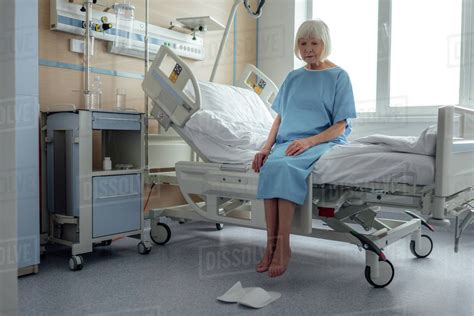 Upset Lonely Senior Woman Sitting On Bed In Hospital Ward Stock Photo