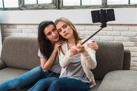 Free Photo Romantic Young Lesbian Couple Sitting On Grey Sofa Taking Selfie With Stick At Home
