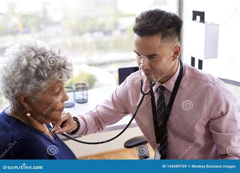Male Doctor In Office Listening To Senior Female Patients Chest Using