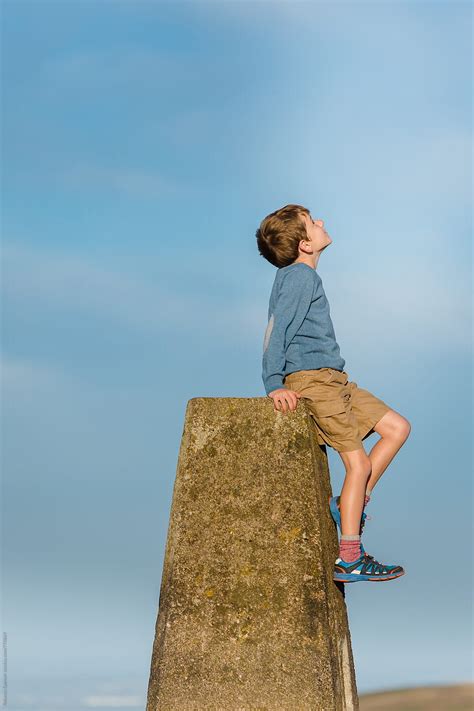 Child Sitting On Trig Point Pillar Looks Up At The Sky By Stocksy