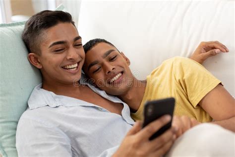 Diverse Gay Male Couple Sitting On Sofa Using Smartphone And Embracing Stock Image Image Of