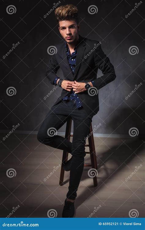 Elegant Guy Posing Seated In Dark Studio Fixing His Jacket Stock Image