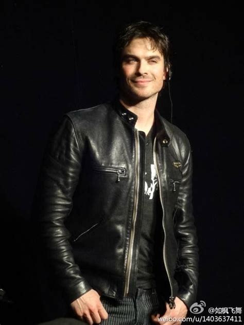 A Man Wearing A Black Leather Jacket Standing In Front Of A Dark Background With His Hands In