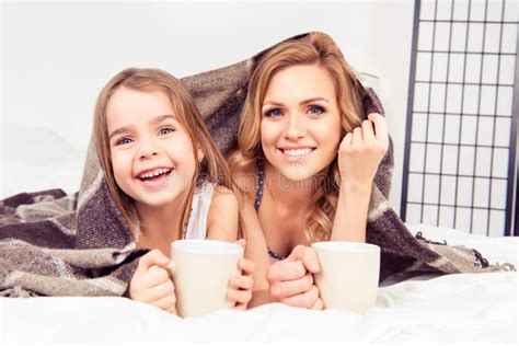 Portrait Of Happy Daughter And Mother Lying Under Plaid With Cups Of