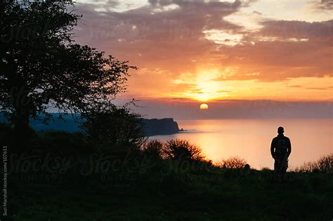 Man Standing On Cliff Watching The Sunset Over The Sea By Suzi Marshall