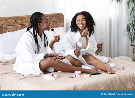 Happy Black Ladies In Bathrobes Sitting On Bed Putting Makeup Stock