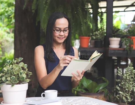 Asian Woman Wearing Eyeglasses Sitting At Table In Garden And Reading