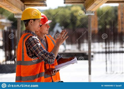 Two Civil Engineers Dressed In Orange Work Vests And Helmets Discuss