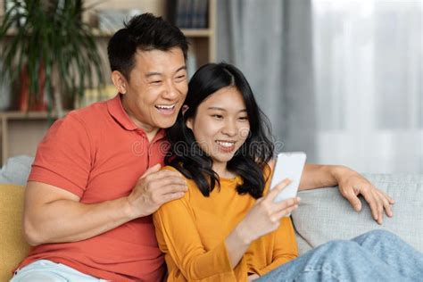 Cheerful Chinese Lovers Using Smartphone Home Interior Stock Image