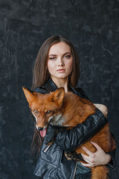 A Woman In Black Jacket Holding A Red Fox On Her Shoulder And Looking
