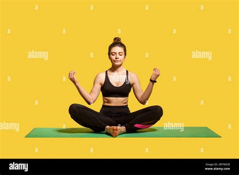 concentrated calm woman with hair bun in tight sportswear sitting in lotus position on gym mat