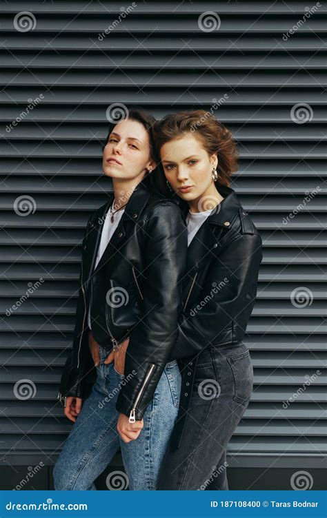 Fashion Portrait Of Two Stylish Girls Standing Against A Black Wall Hugging And Posing At The