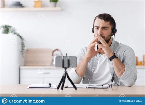 Pensive Mature Caucasian Guy Teacher In Headphones Looks At Smartphone Webcam At Kitchen