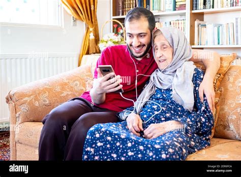 Happy Arabic Muslim Grand Mother And Her Son Sitting Together On Sofa And Using Mobiles Stock