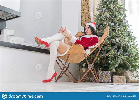 Beautiful Girl In Red Santa Suit Sits On The Floor Near Fireplace And Christmas Tree With A Gift