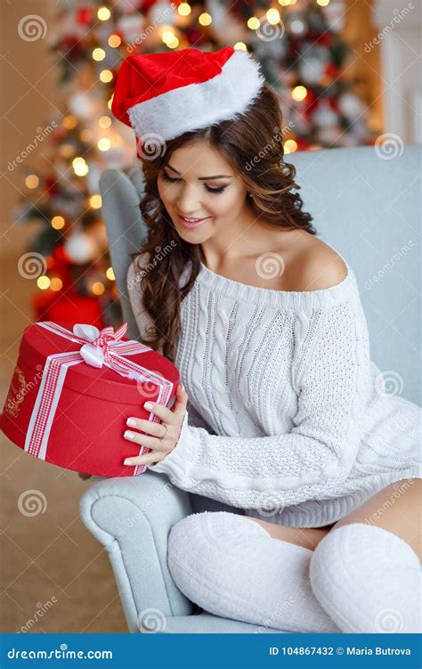 beautiful slender brunette girl in santa hat sits in a cozy armchair smiling and looking at a