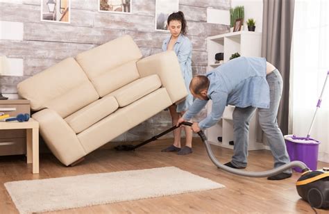 Esposa Levanta El Sofá Mientras Su Esposo Limpia El Polvo Debajo De él Con Una Aspiradora Foto