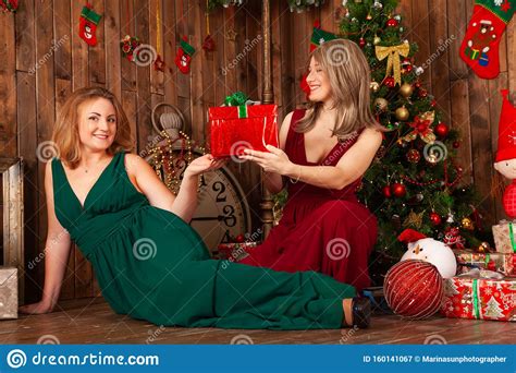 Two Elegant Women In Red And Green Long Evening Dress Posing Near Christmas Tree Indoors Stock