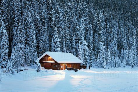 A Snow Covered Log Cabin On A Snow Covered Lakeshore Surrounded By Evergreen Trees At Dusklake