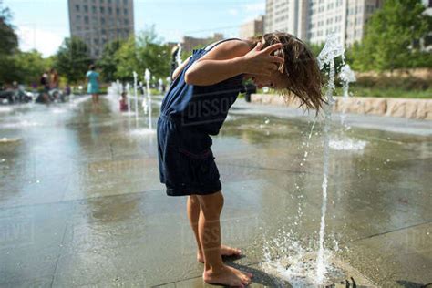 Side View Of Playful Girl Bending Over Fountain In Park During Sunny