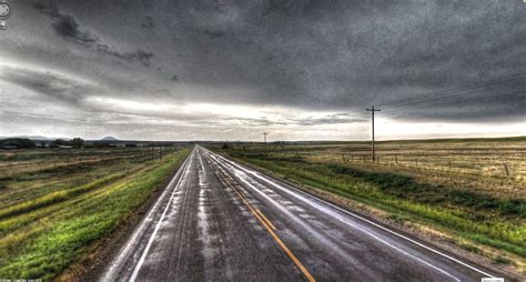 Gray Asphalt Road Between Green Fields Of Grass Under Gray Cloudy Sky