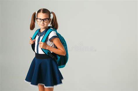 Cute Schoolgirl In Glasses With Blue Backpack Looking At Camera On Grey