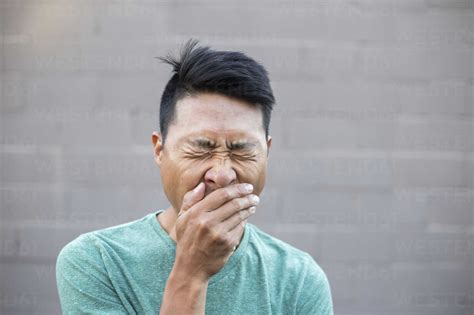 Close Up Of Man With Hands Covering Mouth While Yawning Against Wall