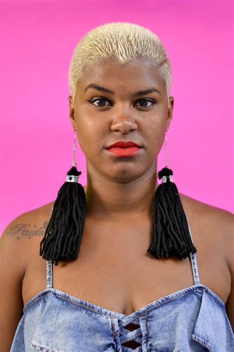 A Woman With Short Blonde Hair Wearing Black Earrings And Denim Overalls Against A Pink Background